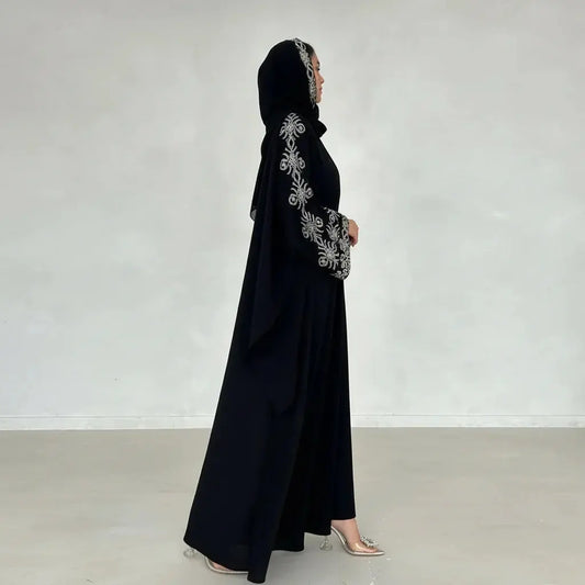 Femme de profil en abaya noire avec broderies argentées sur manches et capuche, chaussée de talons