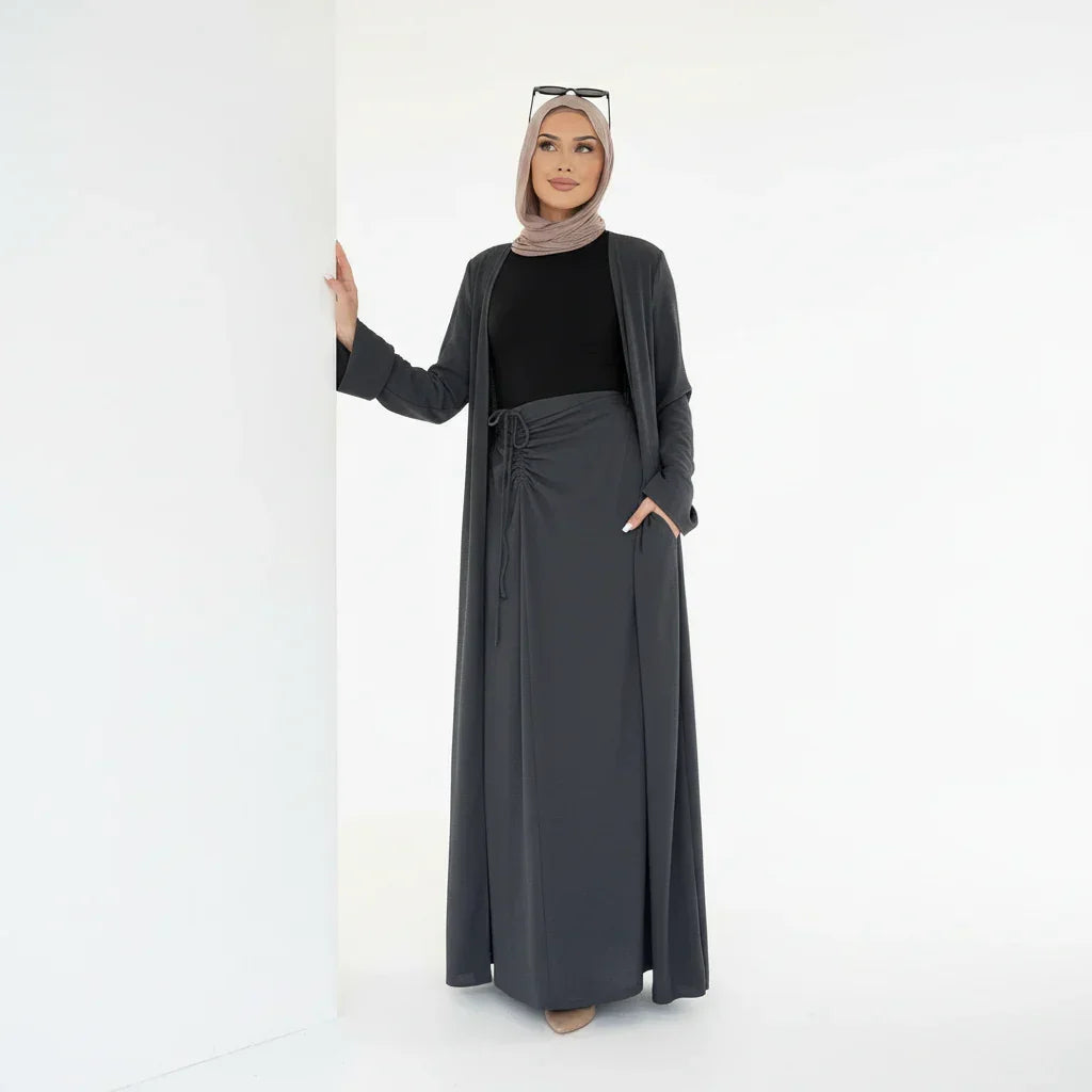 Femme en hijab beige portant une robe longue noire et manteau gris foncé dans un fond blanc