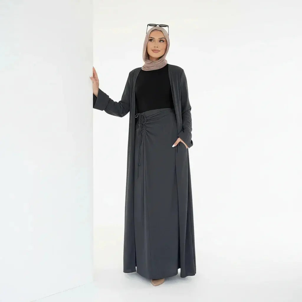 Femme en hijab beige portant une robe longue noire et manteau gris foncé dans un fond blanc