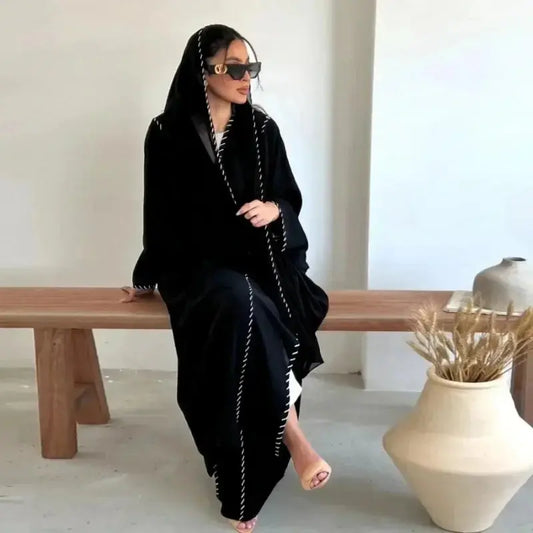 Femme en abaya noire avec coutures blanches assise sur un banc en bois, intérieur minimaliste
