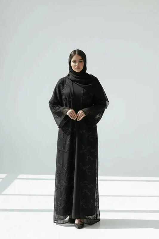 Femme en abaya noire brodée avec hijab noir, debout sur fond blanc minimaliste
